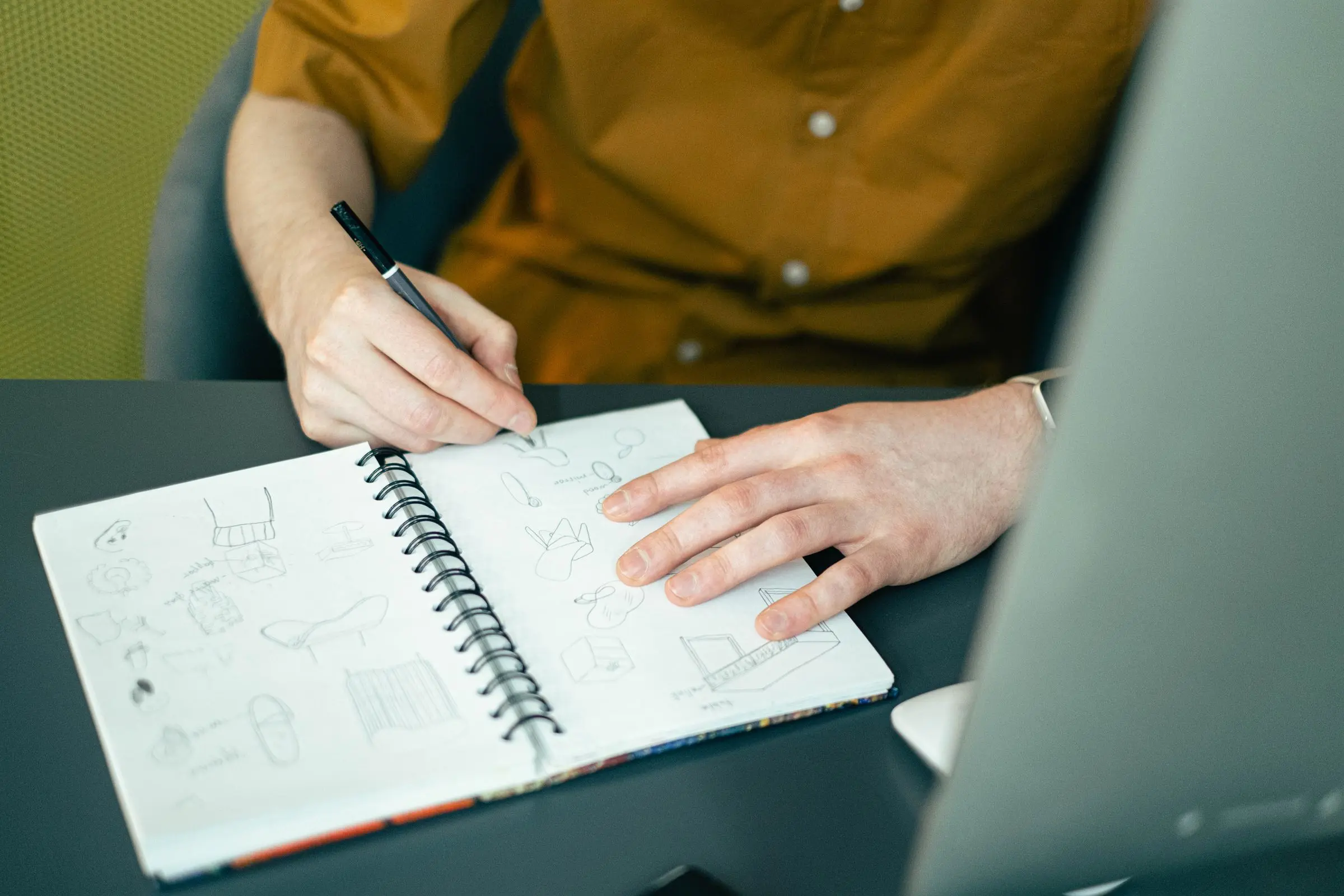 A designer sketching ideas in a notebook beside a laptop